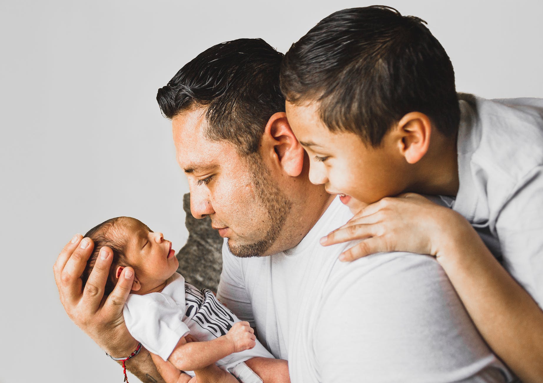 An image of a father with his older looking over his shoulder at the young infant he's holding.